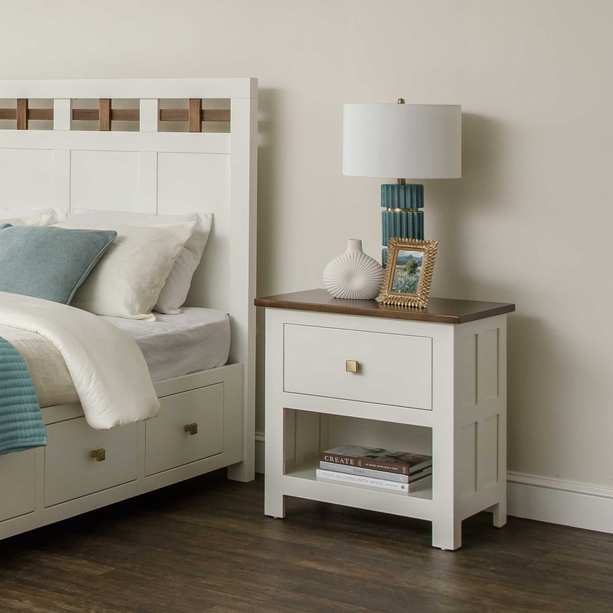 White nightstand with brown top, single drawer and open shelf holding books beside a white lattice storage bed
