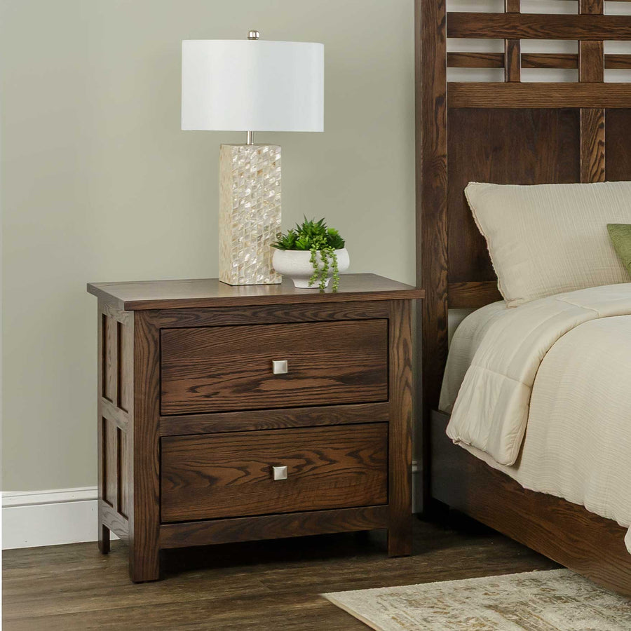 Two-drawer oak nightstand in dark finish next to a bed, topped with a tile lamp and a small potted plant