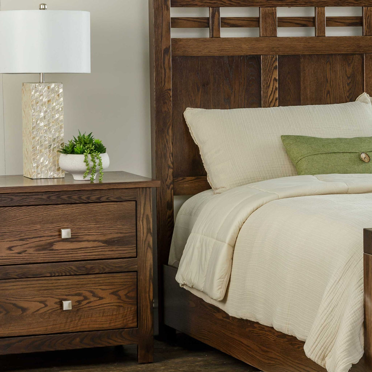 Bedside view of the dark oak two-drawer nightstand and lattice panel headboard with cream and green bedding