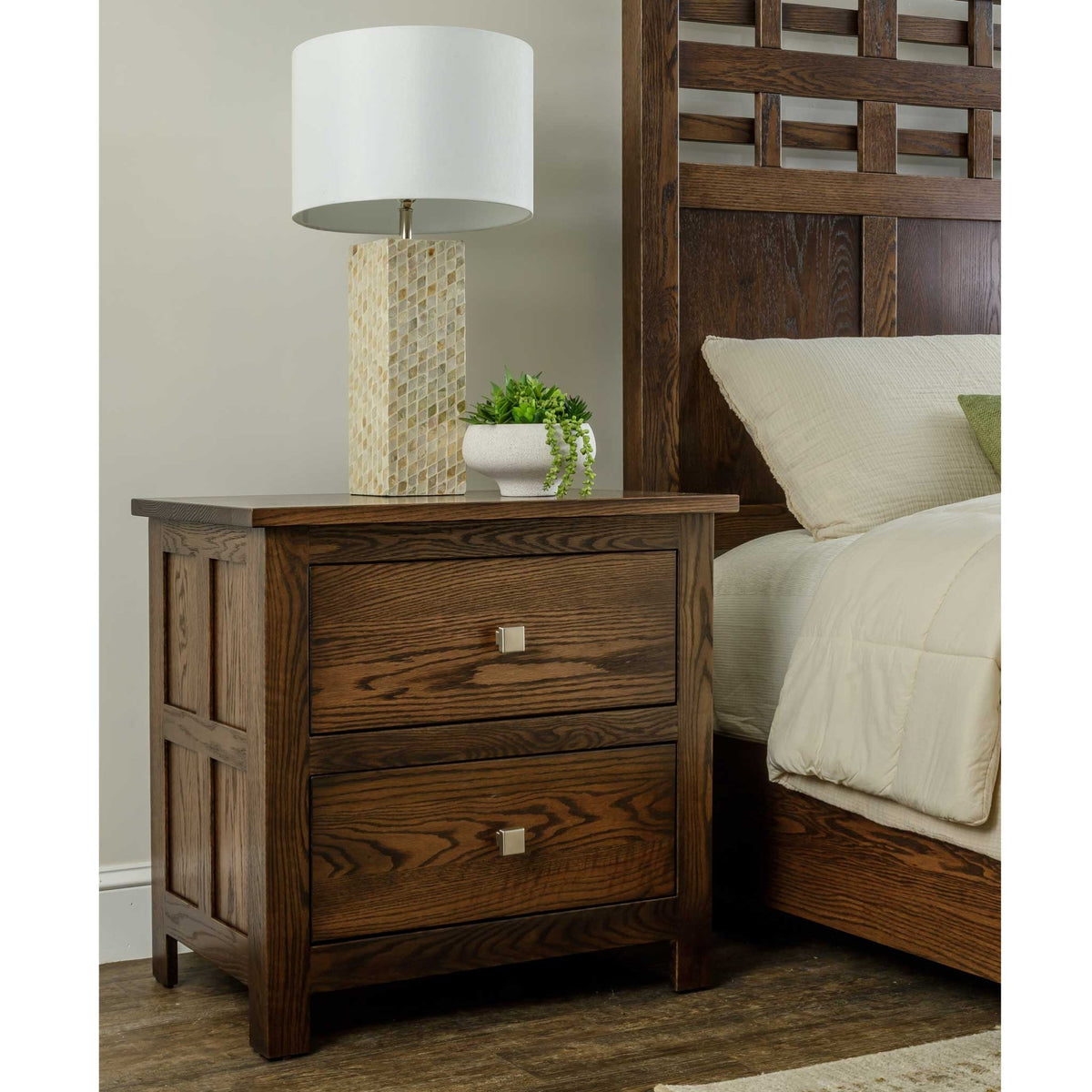 Dark oak two-drawer nightstand with square nickel knobs and paneled sides styled with a lamp beside a lattice bed