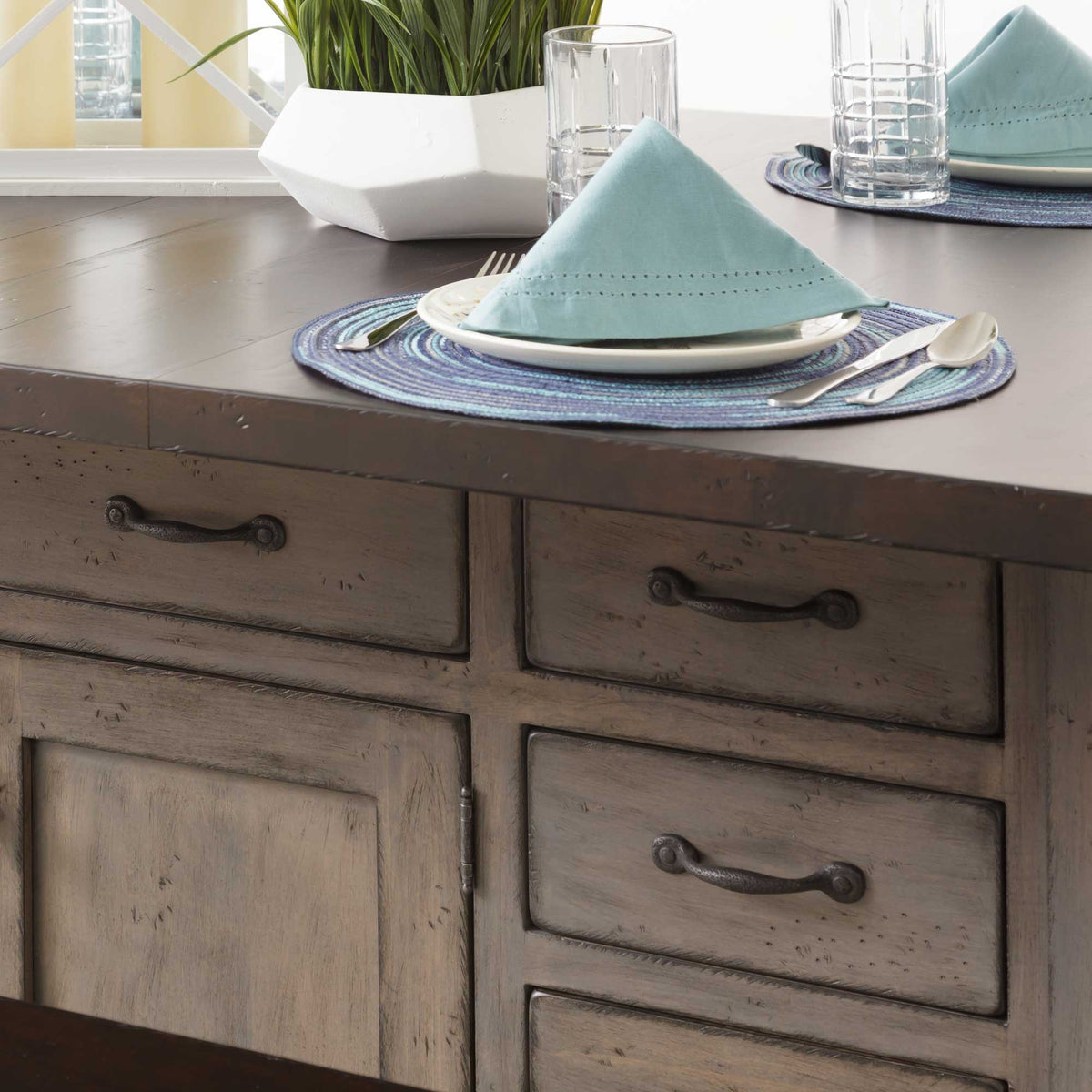 Close view of the weathered gray island cabinet with bronze bin pulls and a dark plank top set with plates and napkins