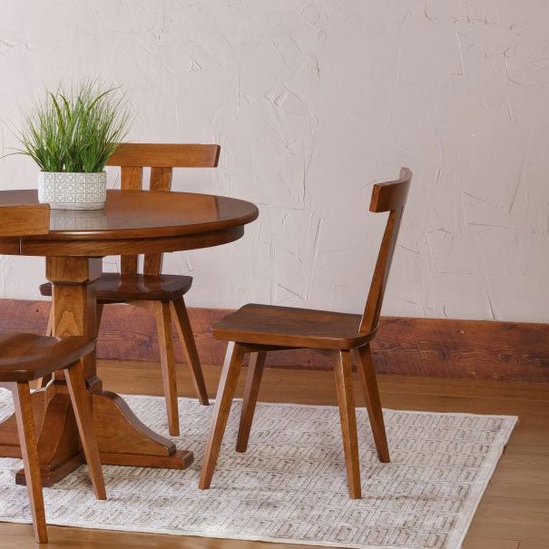 Cherry Astoria dining chairs with T-shaped backs around a round pedestal table on a woven rug by a plaster wall