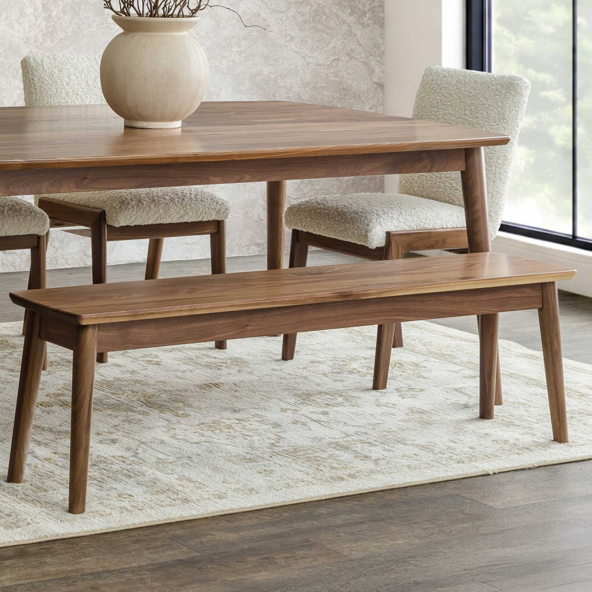 Walnut dining bench with rounded plank seat and tapered legs beside a matching table and boucle chairs