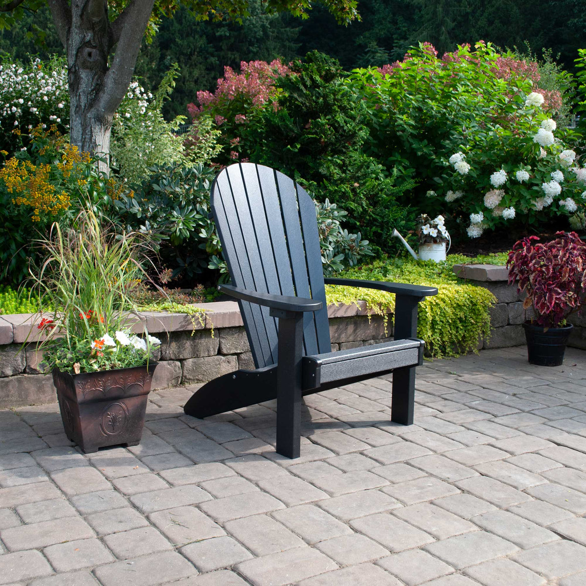 Black poly Adirondack chair with fanned slat back and wide arms on a paver patio surrounded by flowering shrubs