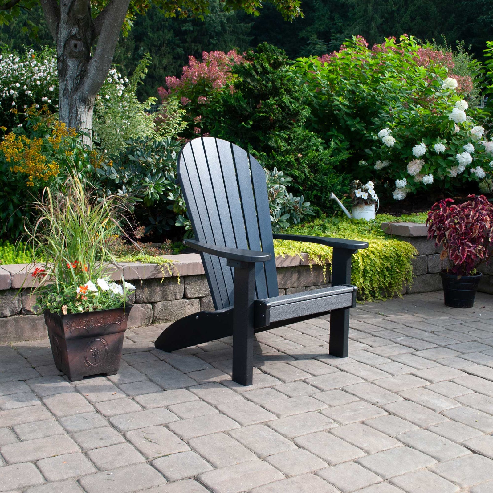 Black poly Adirondack chair with fanned slat back and wide arms on a paver patio surrounded by flowering shrubs