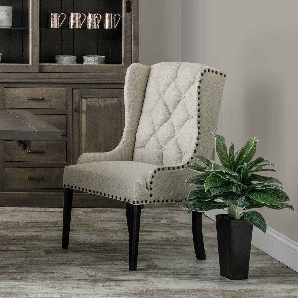 Beige tufted wingback host chair with dark nailhead trim and black legs styled beside a gray hutch with copper mugs