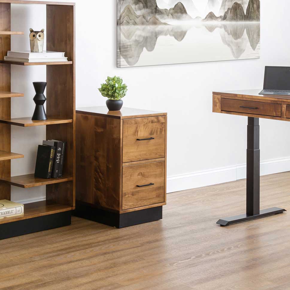 Modern two drawer file cabinet in golden alder with black bar pulls beside a lift desk and open shelf bookcase