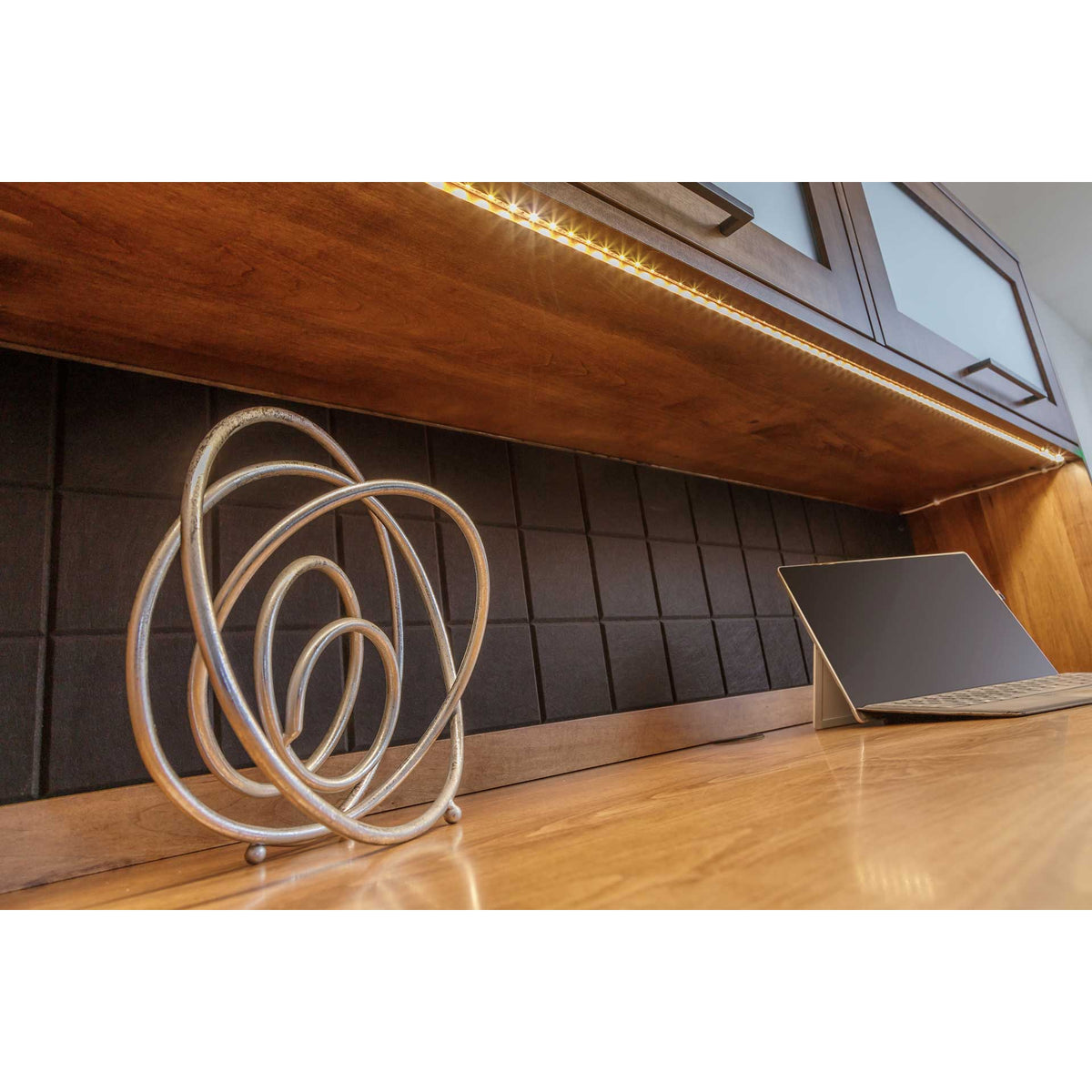 Warm lit desktop under the hutch with a strip light, tablet and looping metal sculpture on the dark tackboard back