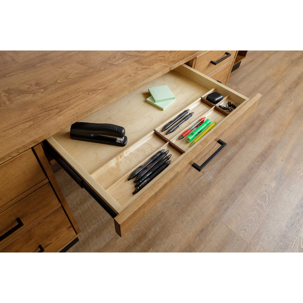 Open center drawer with maple divided tray holding a stapler, pens, highlighters and sticky notes in a modern wood desk