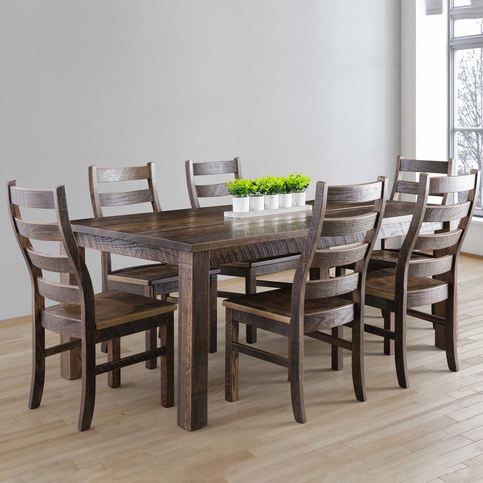 Brown weathered farm table with six ladder back chairs and a small planter centerpiece in a sunlit room
