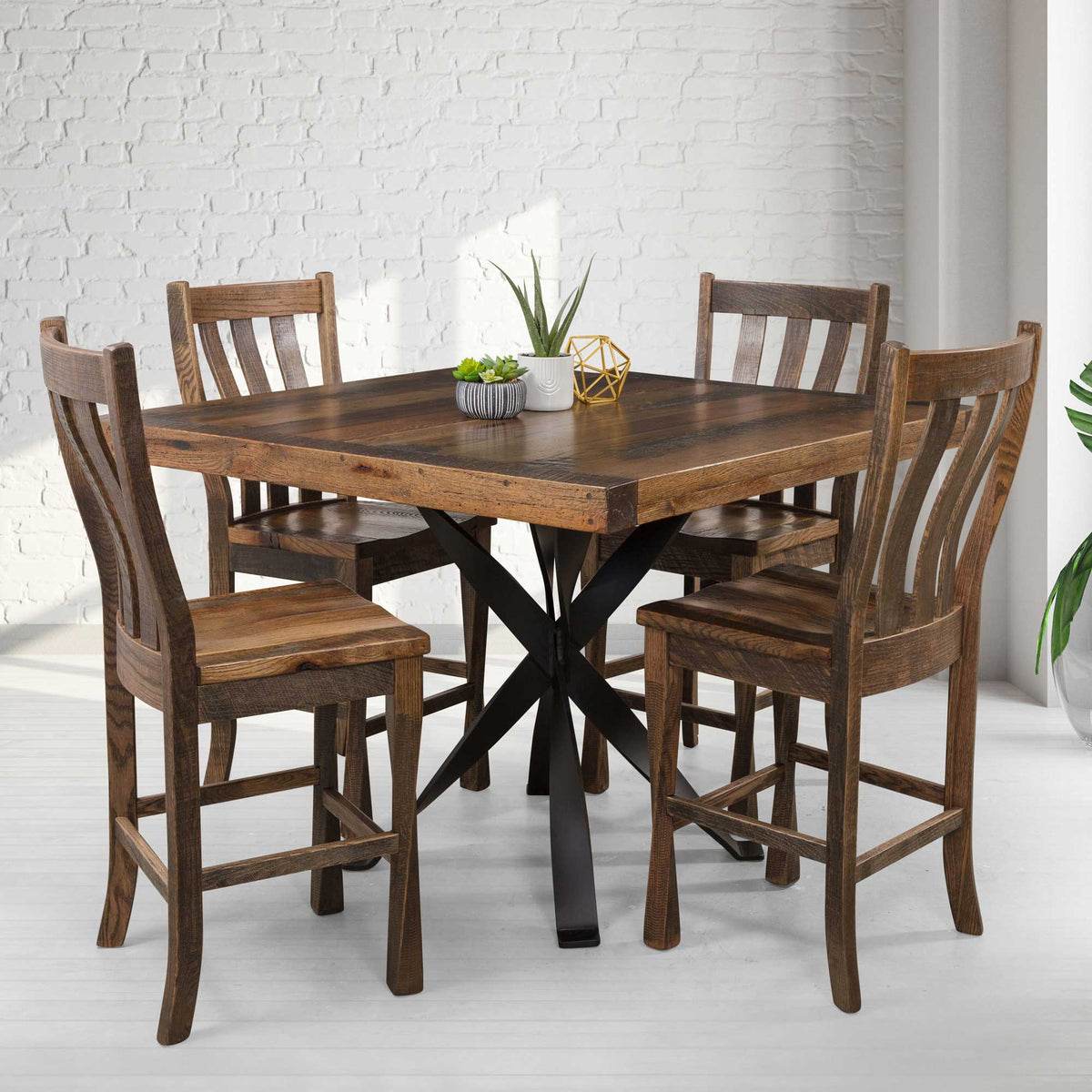 Square rustic counter height table with a black iron X base and four tall slat back stools in a white brick room