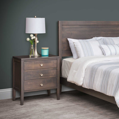 Dark gray brown three drawer bedside chest with round knobs, styled with a lamp next to a plank headboard bed