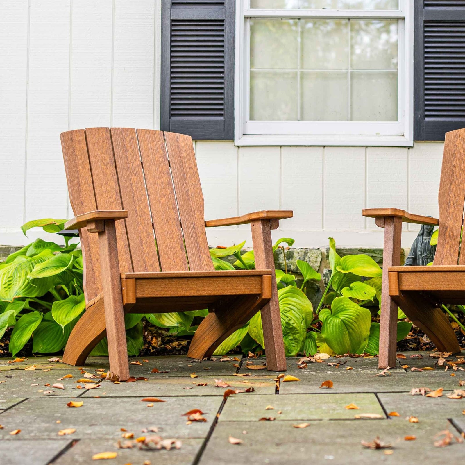 Two antique mahogany poly Adirondack chairs with squared modern arms in front of a white house with hostas