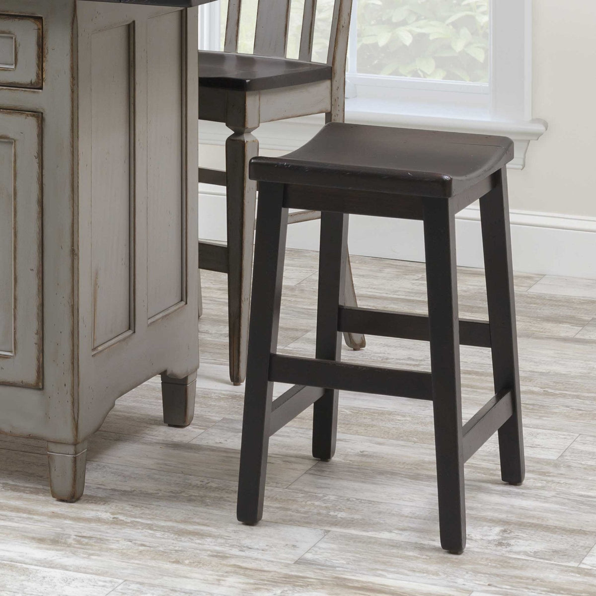 Black painted saddle seat bar stool with tapered legs and side stretchers beside a distressed gray kitchen island