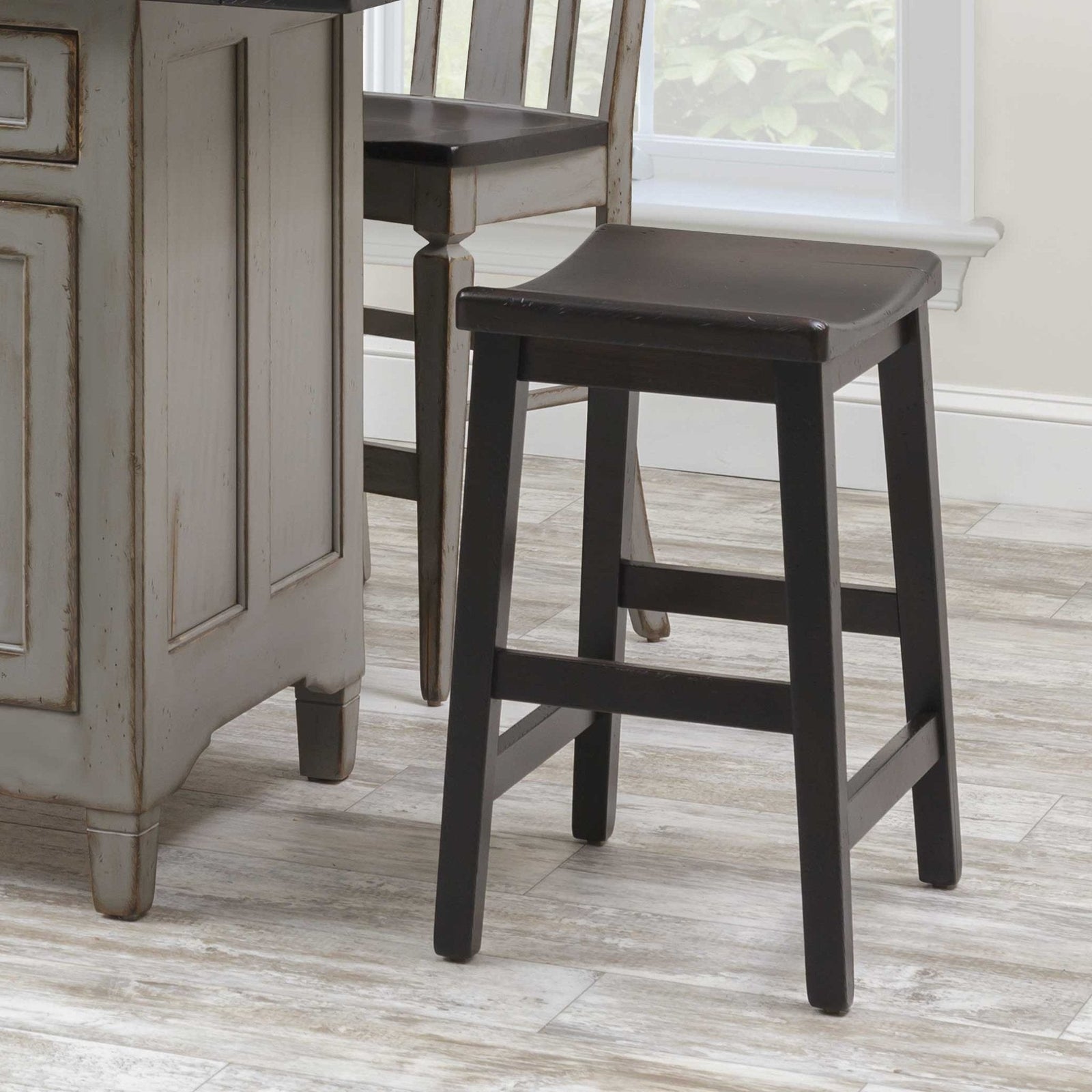 Black painted saddle seat bar stool with tapered legs and side stretchers beside a distressed gray kitchen island