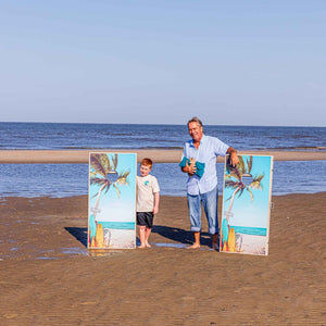 Man and boy standing on the beach beside two upright palm tree print cornhole boards