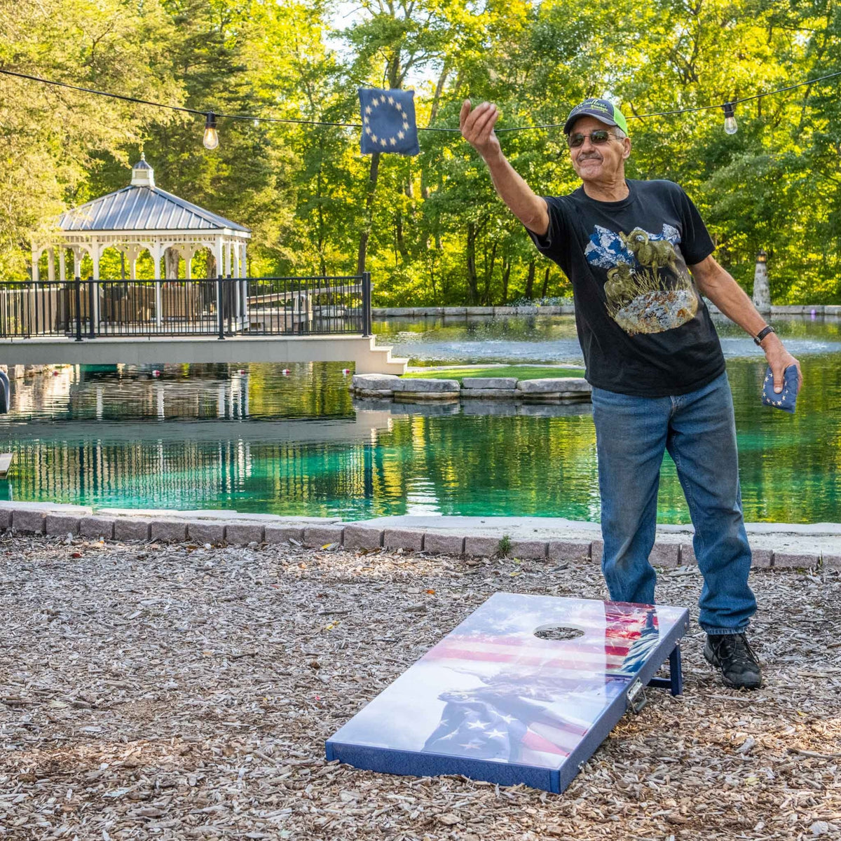 Player tossing a bean bag toward a patriotic flag and eagle cornhole board in a landscaped yard