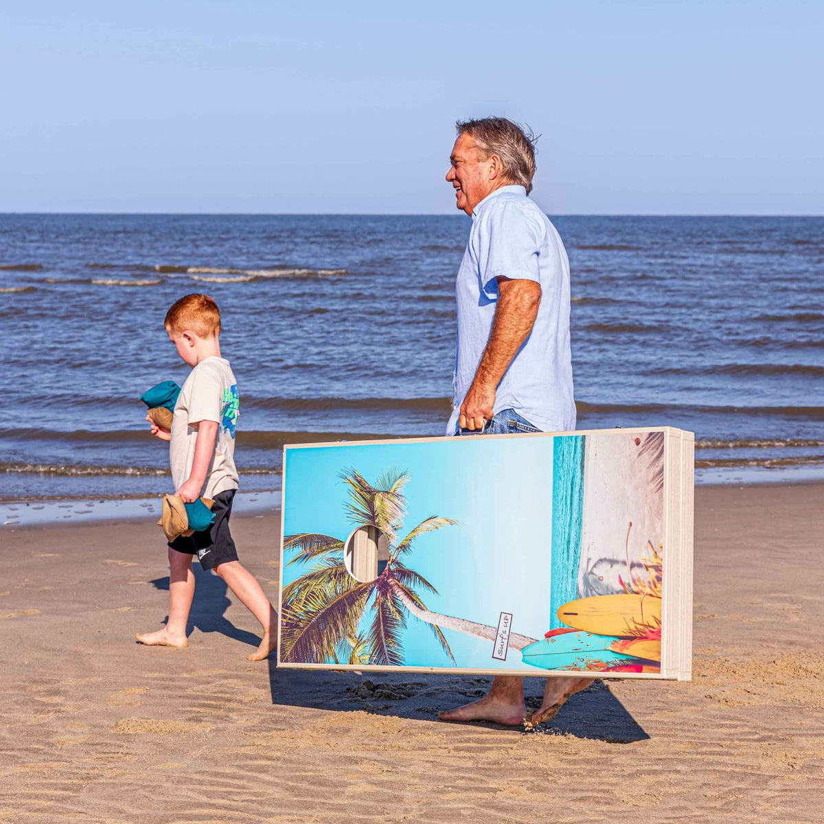 Man and boy carrying a palm tree and surfboard printed cornhole board across the beach
