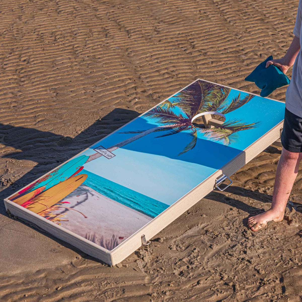 Single palm tree cornhole board on the beach with a player holding a teal bean bag