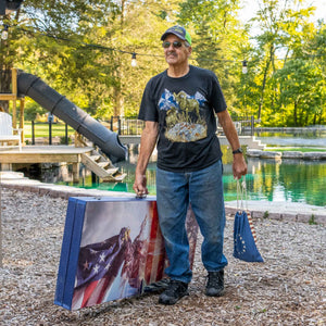 Man holding a folded cornhole board printed with an American flag and eagle design beside a backyard pond