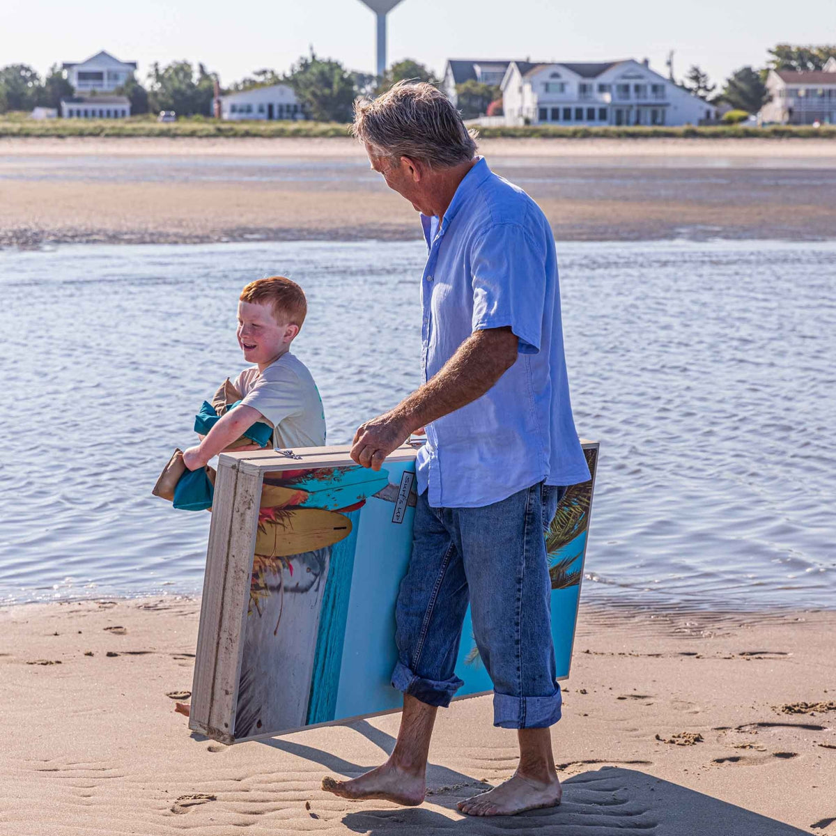Grandfather and grandson carrying folded tropical print cornhole boards along the shoreline