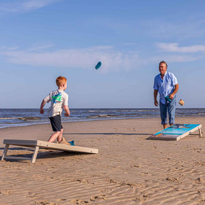 Boy and man playing cornhole on the beach with bean bags in mid air between two boards