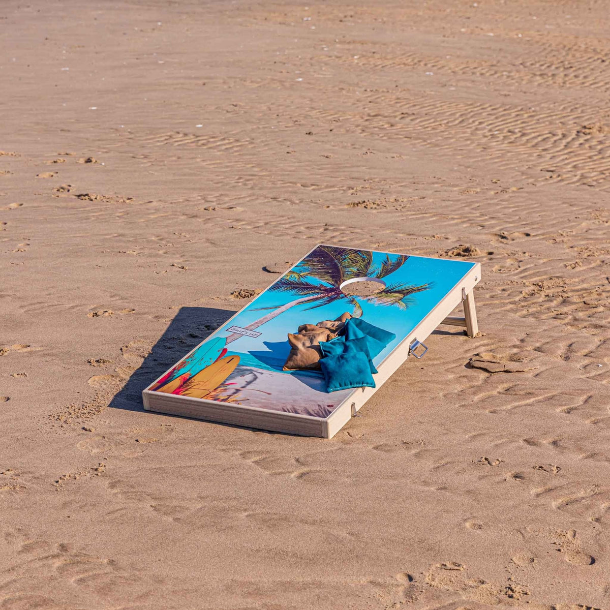 Tropical palm tree cornhole board on the sand with tan and teal bean bags resting on top