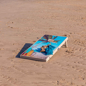 Tropical palm tree cornhole board on the sand with tan and teal bean bags resting on top