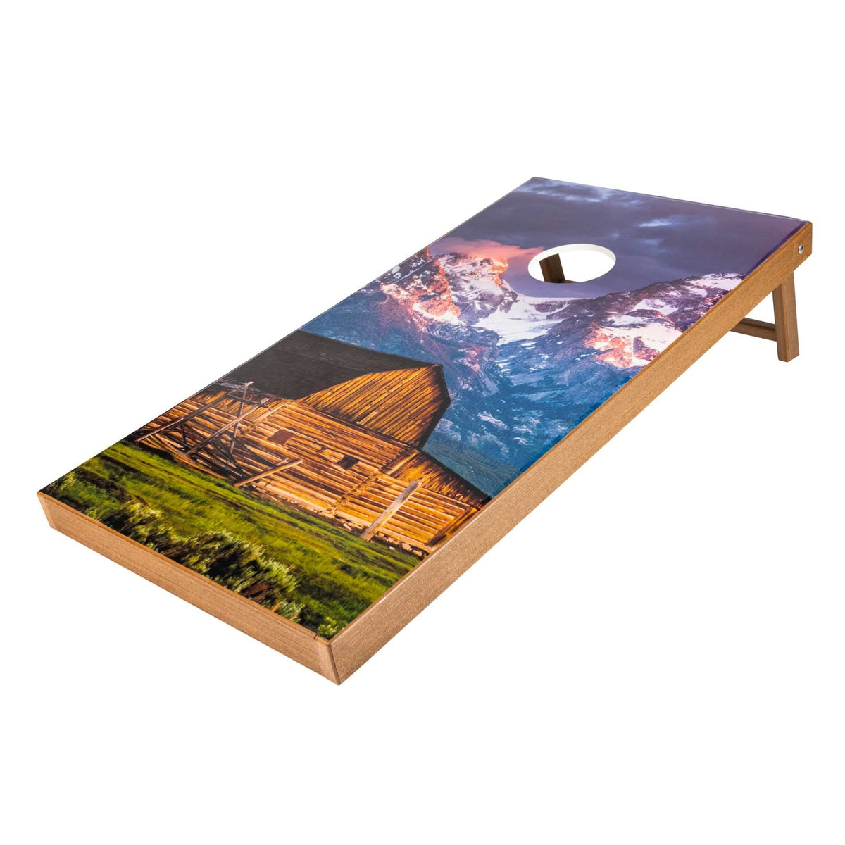 Cornhole board printed with a mountain sunrise and rustic log barn, natural wood frame and folding legs