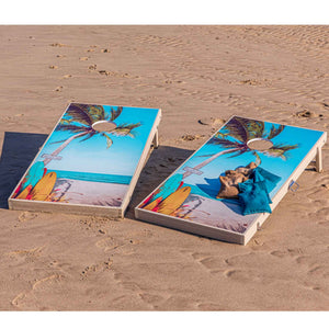 Pair of beach themed cornhole boards with palm tree graphics and teal bean bags set up on the sand