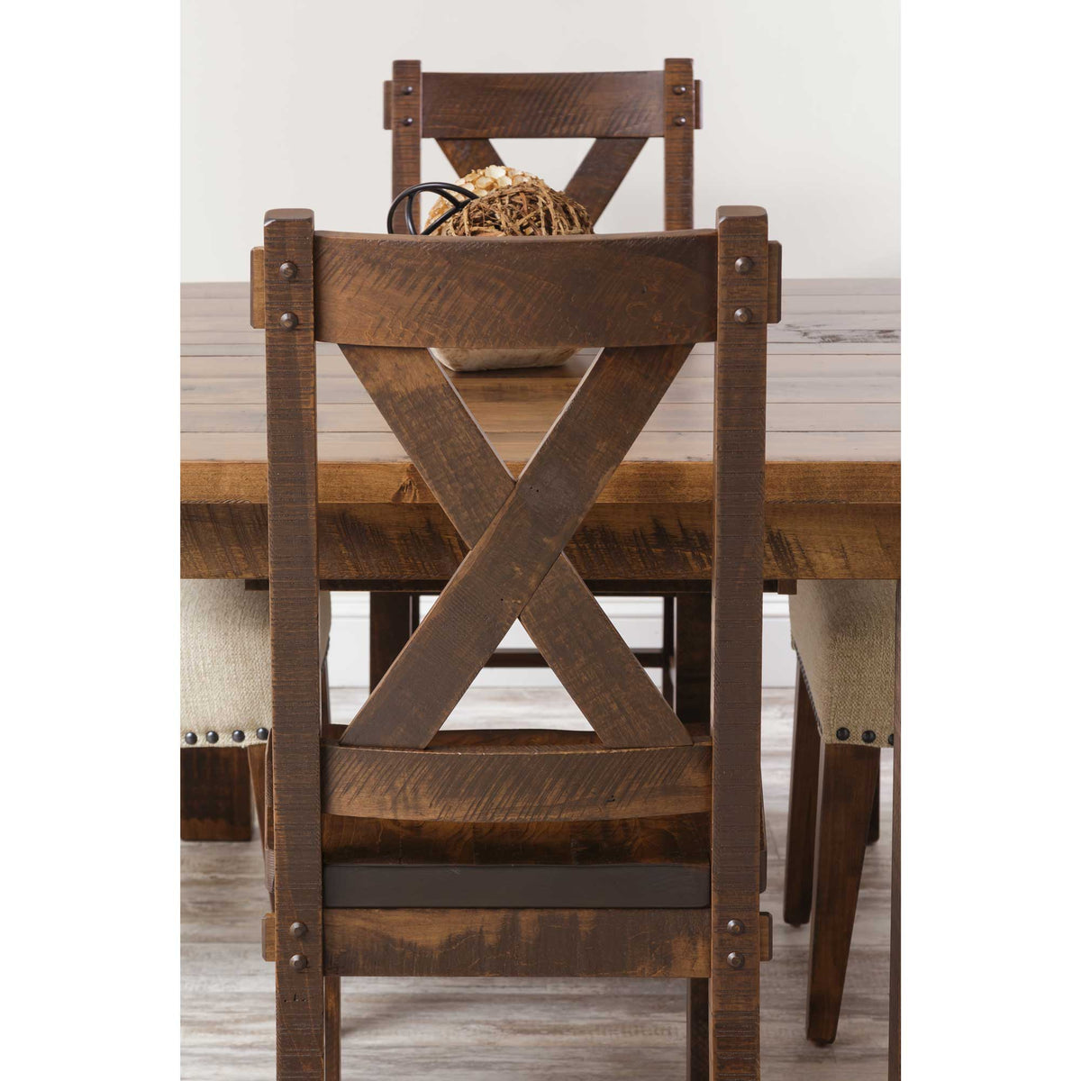 Back view of two rustic X back chairs at a rough-sawn plank dining table with nailhead upholstered chairs