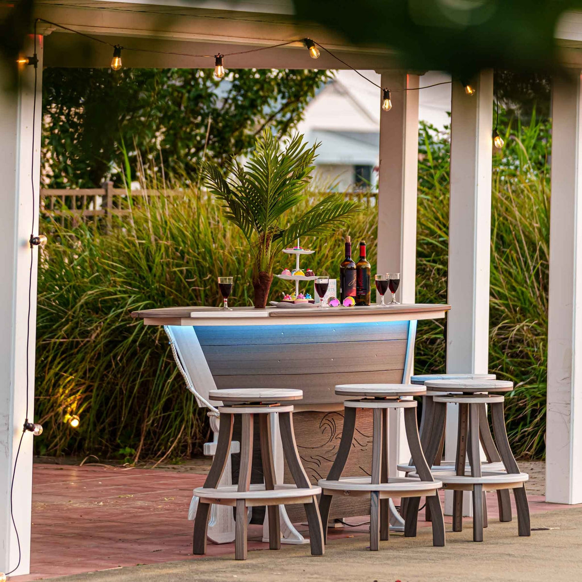 Boat shaped outdoor bar lit blue under a pergola with string lights, palm and six stools