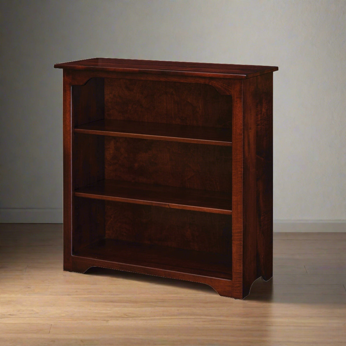 Three-shelf bookcase in rich cherry stain standing against a gray wall on light wood flooring
