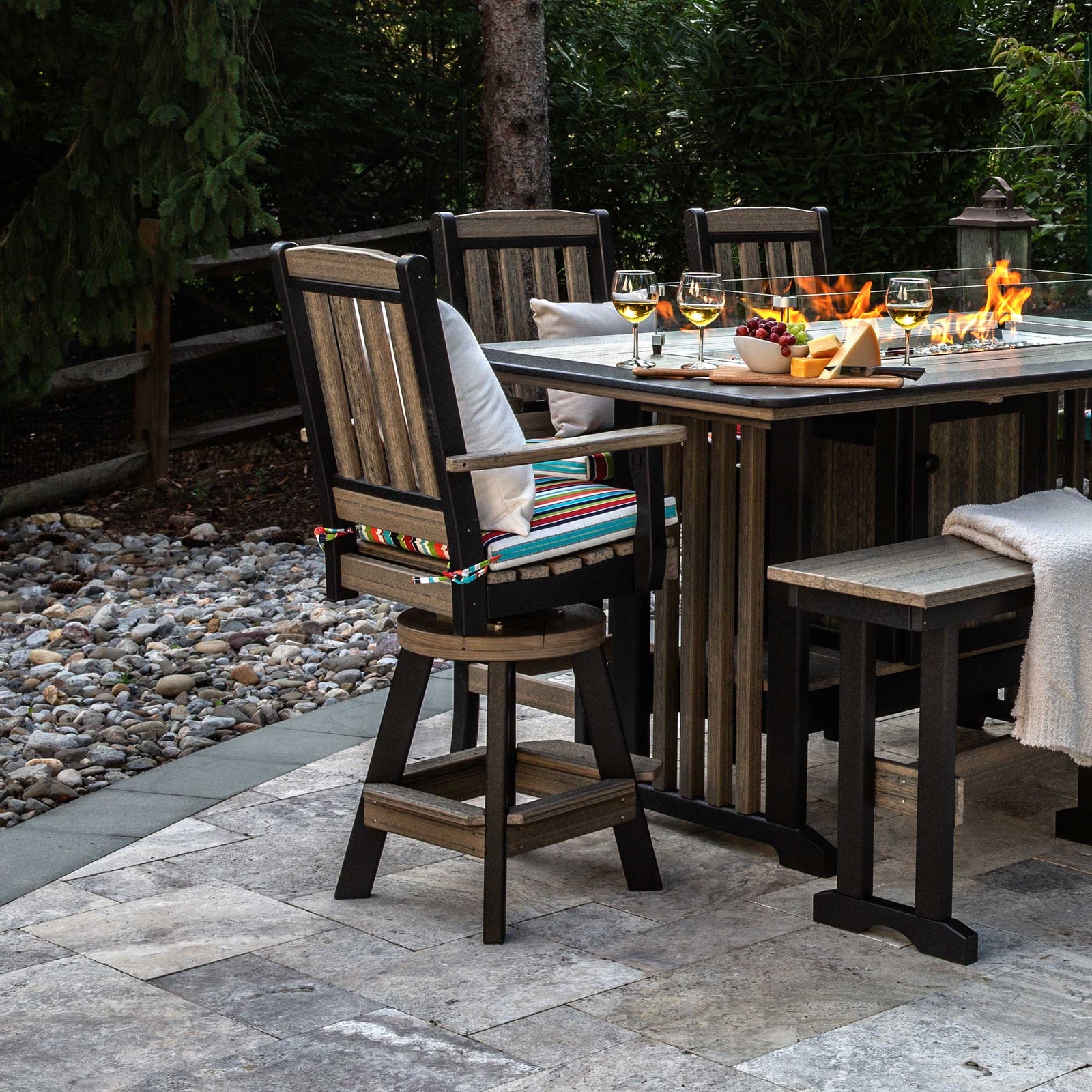 Driftwood and black poly swivel bar chairs with striped cushions around a lit fire pit dining table on a stone patio