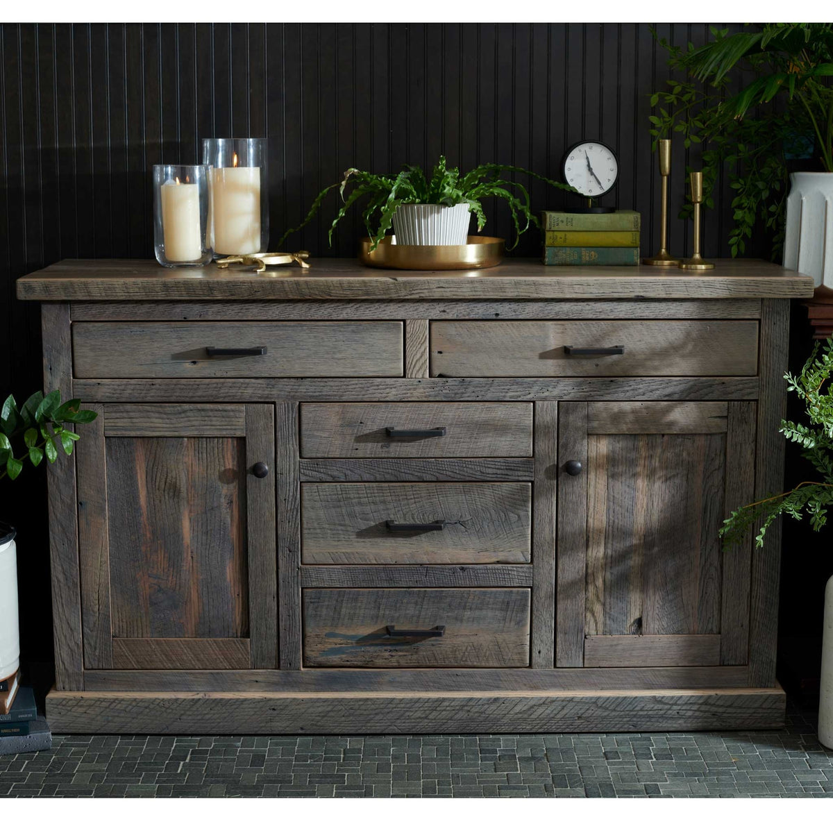 Weathered gray barnwood buffet with black hardware displaying candles, brass accents, books and a potted fern
