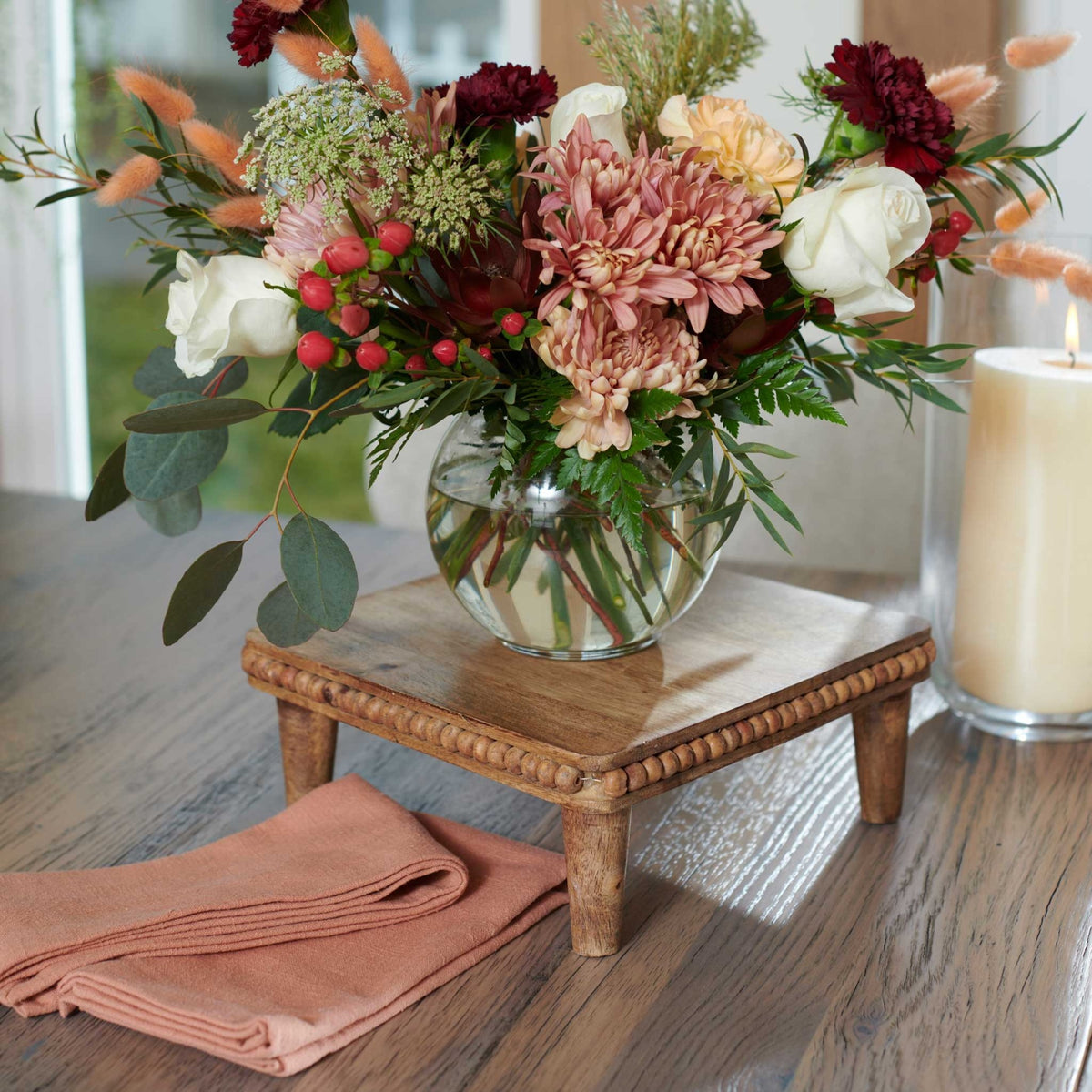 Fall bouquet in a round glass vase on a beaded wood riser sitting on the weathered gray reclaimed wood dining table