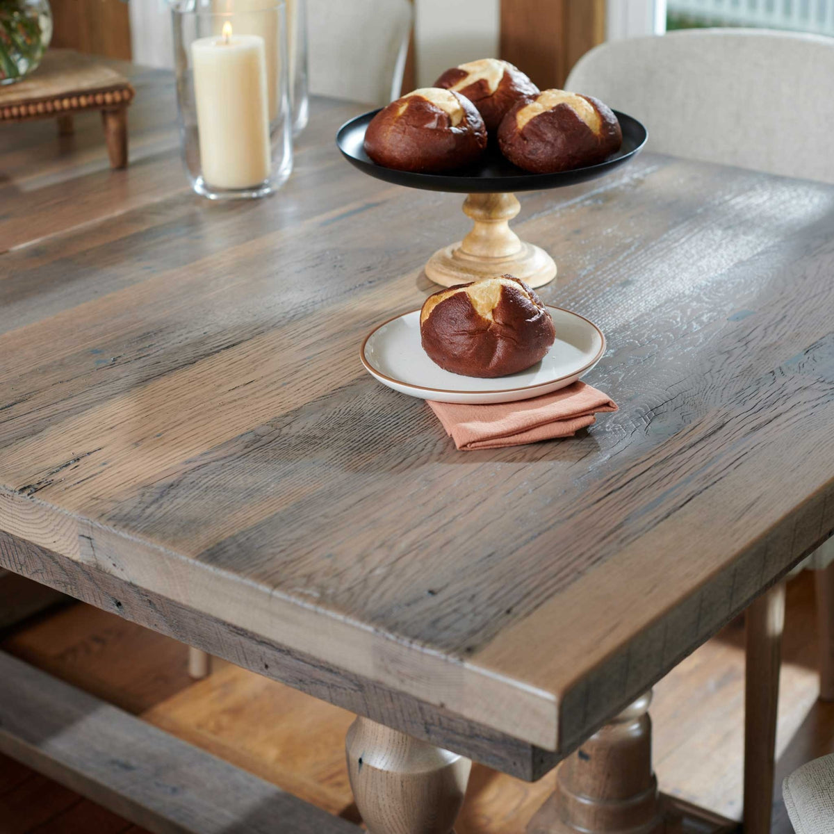 Rough-sawn gray reclaimed table top styled with pretzel rolls on a black pedestal stand and a rust linen napkin