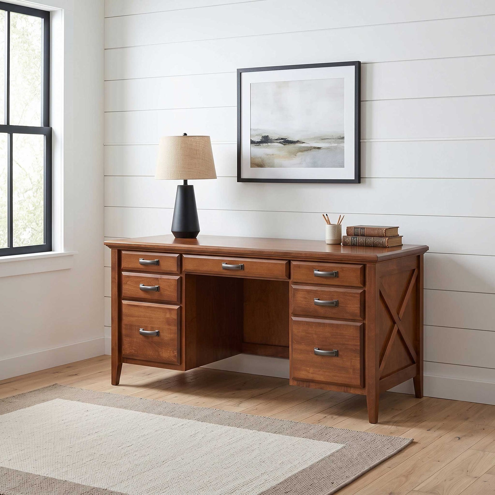 A mid-century modern writing desk in warm medium brown wood, styled in a bright home office with a black base lamp, framed wall art, and a light wood floor.