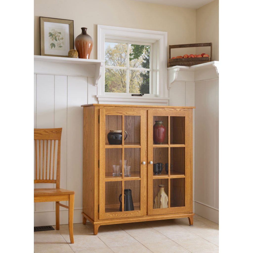 Oak display cabinet with two mullioned glass doors and three shelves of pottery in a white paneled kitchen