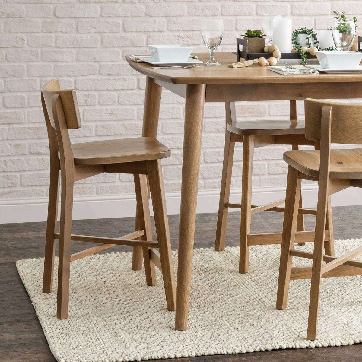 Natural oak counter stools with curved back rests and saddle seats at a light oak table by a white brick wall