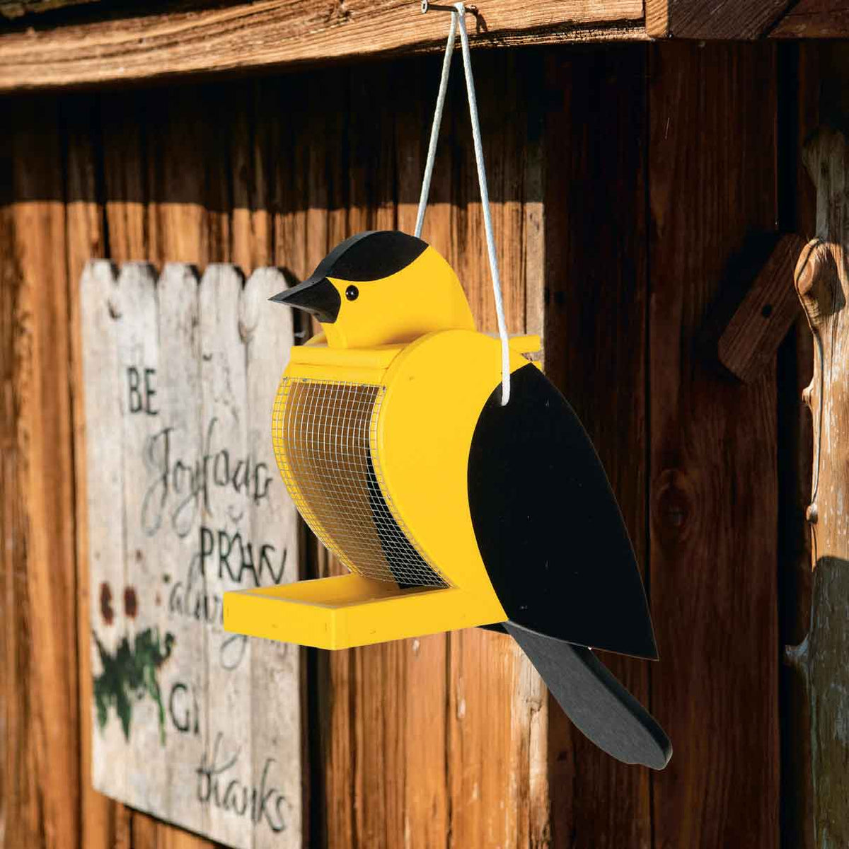 Yellow and black goldfinch bird feeder filled with seed, hanging on a rustic barn wood wall