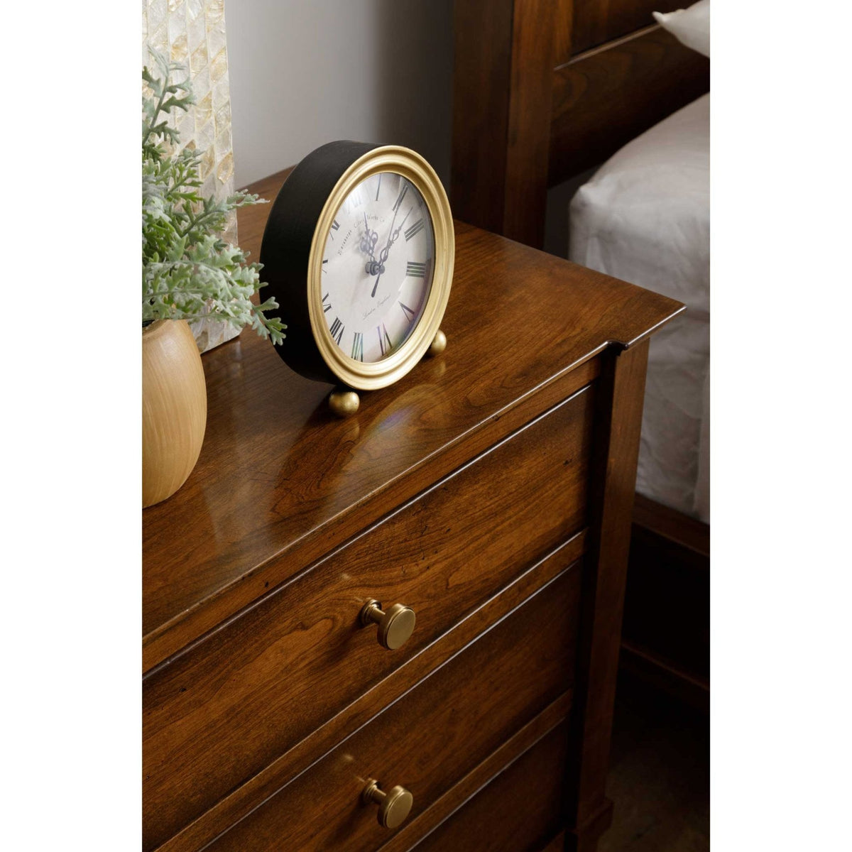 Brass framed clock and greenery on a cherry nightstand top beside a bed with white bedding