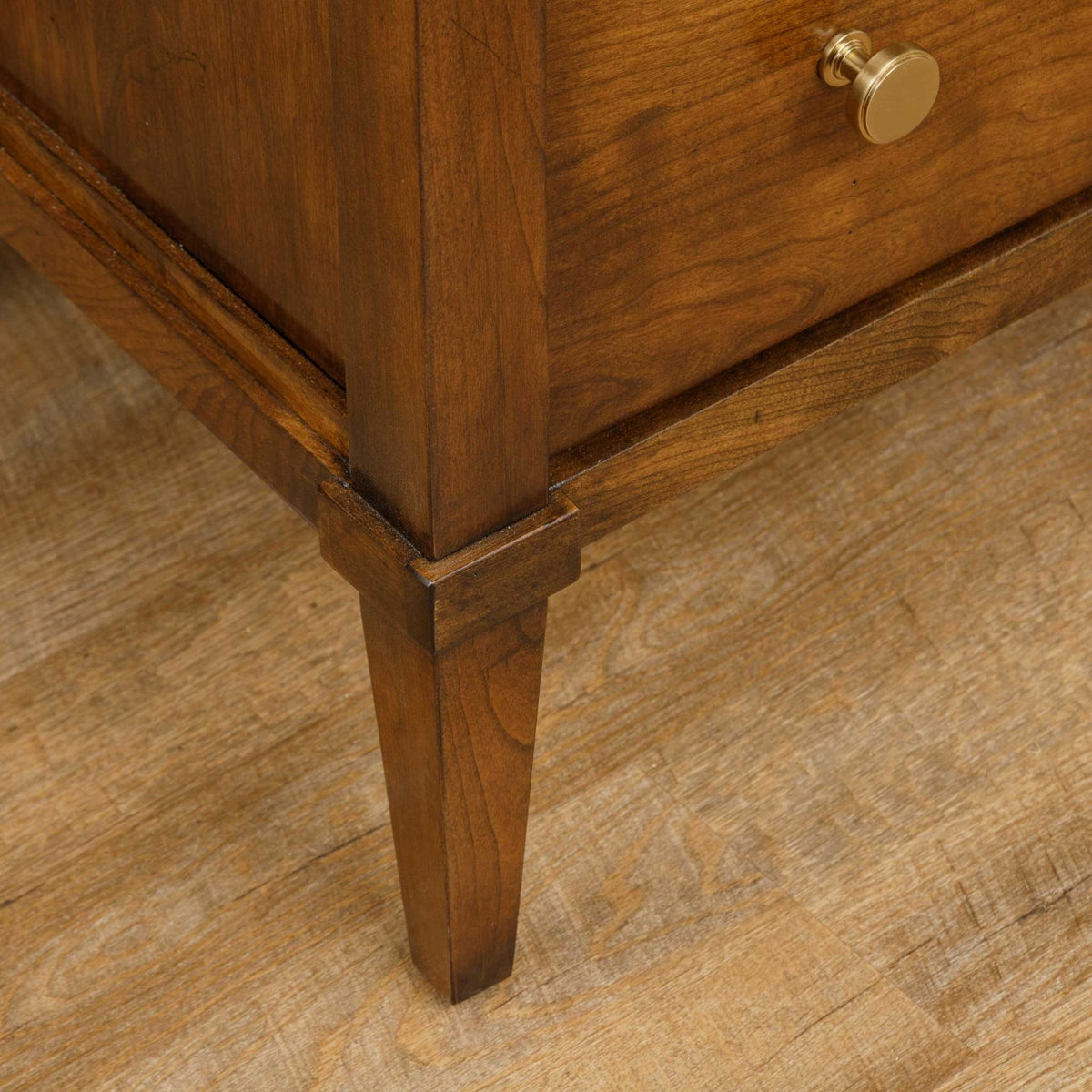 Base of the cherry nightstand with square tapered leg, apron molding and lower drawer knob on wood flooring