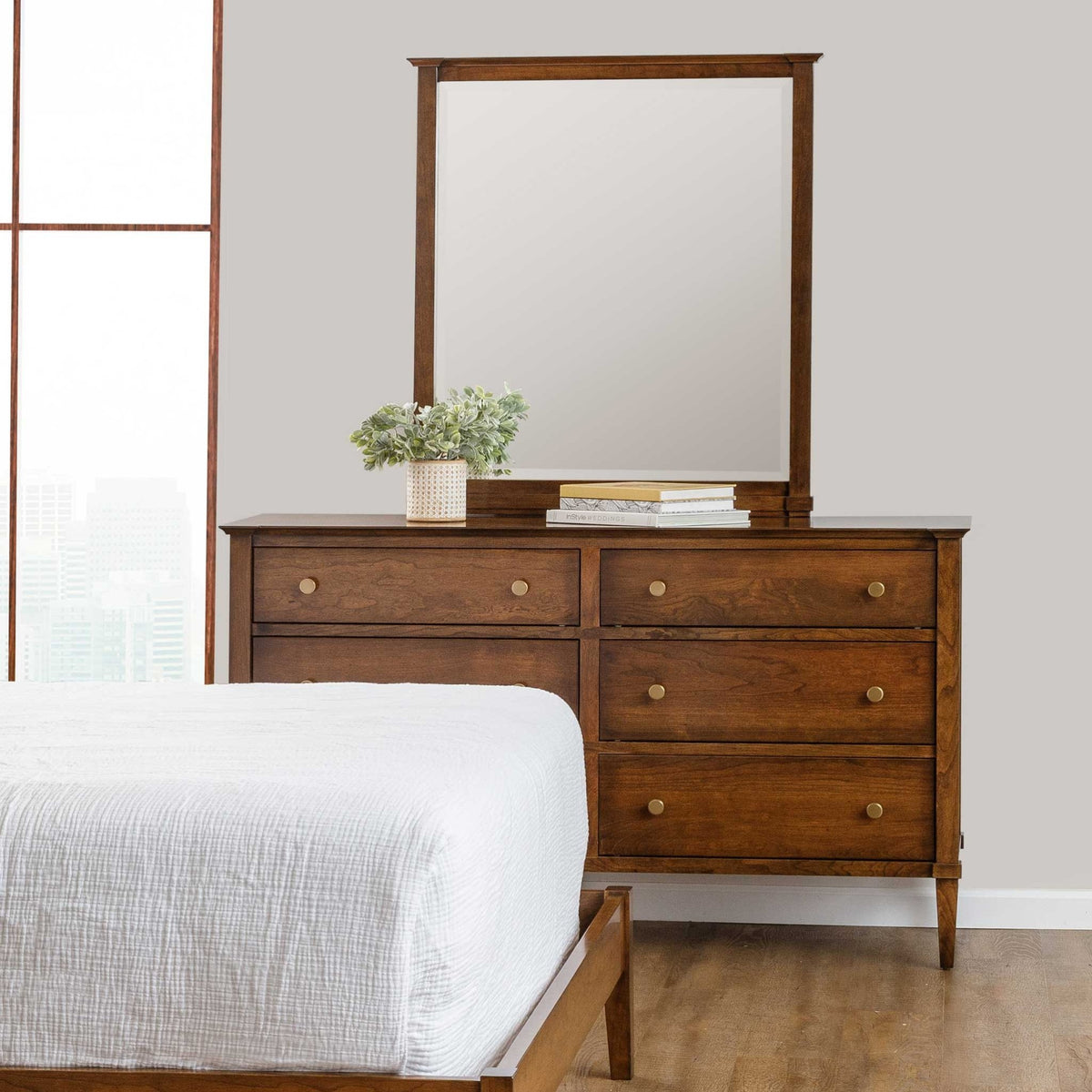 Cherry six drawer triple dresser with a tall mirror and greenery, next to a bed by a bright window