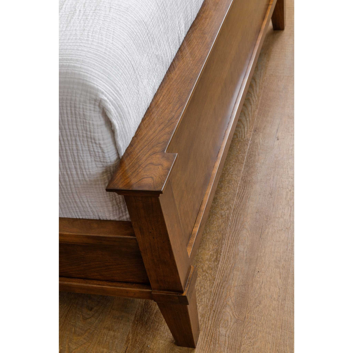 Angled view of a cherry bed footboard panel with notched rail detail beside white bedding on wood flooring