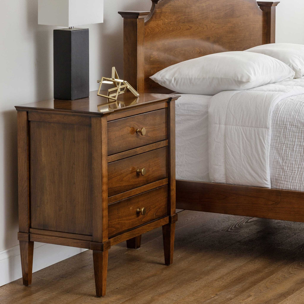 Three-drawer nightstand in warm cherry next to an arched panel bed, topped with a black lamp and brass geometric decor