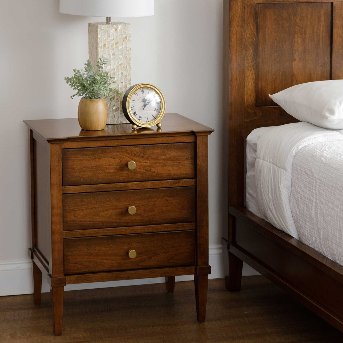 Cherry nightstand at bedside holding a mother-of-pearl lamp, brass alarm clock and a small vase of greenery