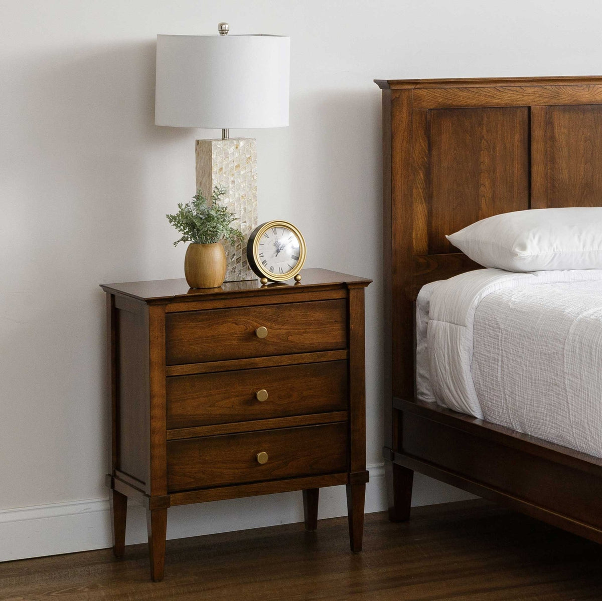 Three-drawer cherry nightstand beside a panel bed, styled with a white shade lamp, brass clock and small potted greenery