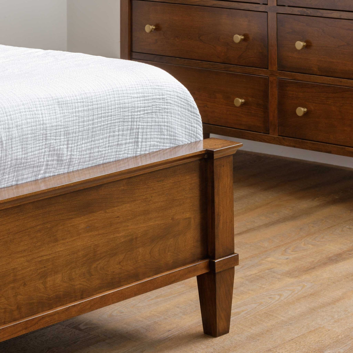 Footboard corner of the cherry panel bed showing the capped post, tapered leg and a dresser with brass knobs behind