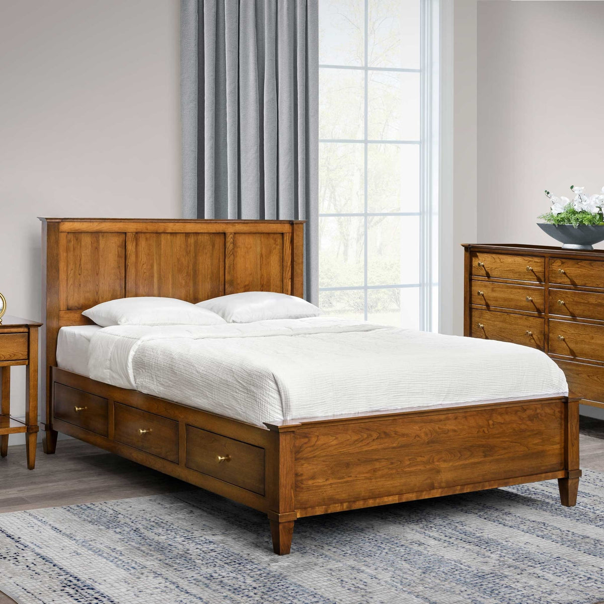 Cherry panel storage bed with three underbed drawers styled in a bright bedroom with gray drapes and a dresser